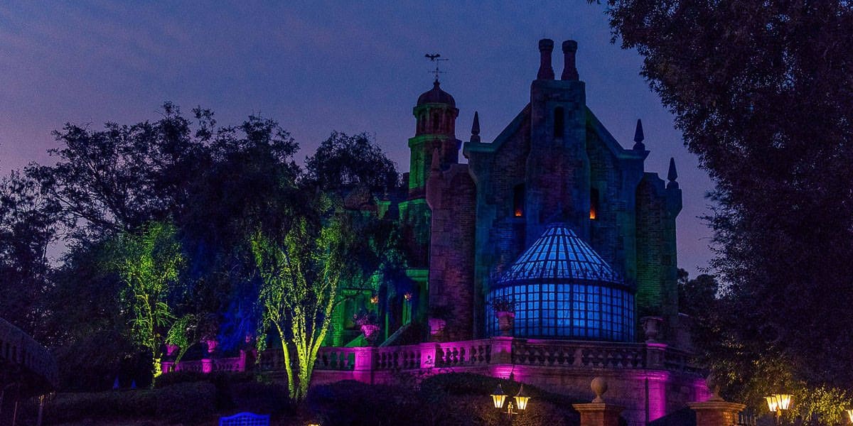 A spooky, dark mansion is lit with eerie green, purple, and blue lights against a twilight sky. Surrounded by trees, the building evokes Disney's Haunted Mansion ride with its distinct glass conservatory, towers, and chimney stacks—perfect for a haunted mansion trivia night.
