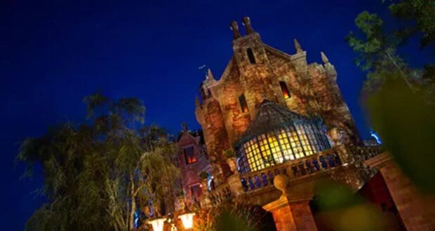 A gothic-style mansion with ornate architecture and glowing windows is illuminated against a dark night sky. The building, which holds Disney's Haunted Mansion ride, is surrounded by trees, adding to the eerie atmosphere.