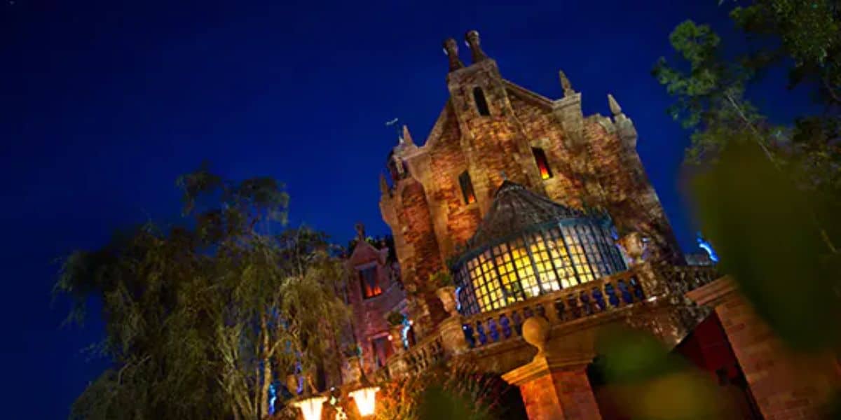 A gothic-style mansion with ornate architecture and glowing windows is illuminated against a dark night sky. The building, which holds Disney's Haunted Mansion ride, is surrounded by trees, adding to the eerie atmosphere.