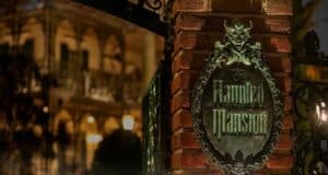 A nighttime image of a brick pillar with an ornate metal sign reading "Haunted Mansion," featuring a demonic face at the top. The background shows a dimly lit, gothic-style building with intricate ironwork, adding to the eerie atmosphere reminiscent of Disney's Haunted Mansion ride.