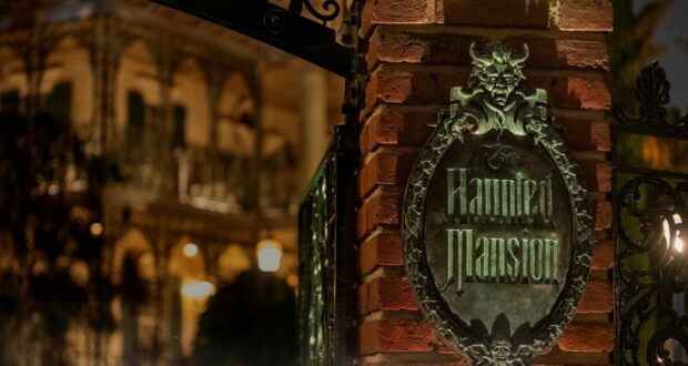 A nighttime image of a brick pillar with an ornate metal sign reading "Haunted Mansion," featuring a demonic face at the top. The background shows a dimly lit, gothic-style building with intricate ironwork, adding to the eerie atmosphere reminiscent of Disney's Haunted Mansion ride.