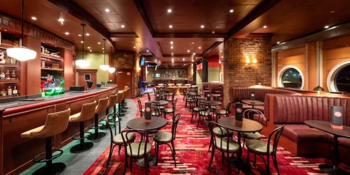 A warmly lit restaurant with a brick wall and wooden ceiling, reminiscent of the Disney Magic Cruise Ship Refurbishment, features a long bar counter with barstools on the left. On the right, round tables surrounded by chairs and booths are set on red and black patterned carpet.