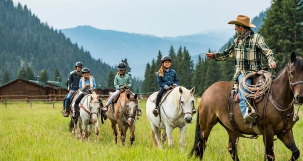 A group of people enjoy a thrilling horseback ride through a lush green field with mountains in the backdrop, guided by an expert. They're equipped with helmets and casual clothing, just like an adventure from New Vacations by Disney. In the distance, a charming wooden cabin can be seen.