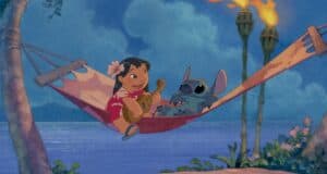 A girl and a blue alien creature are lounging on a hammock by the ocean, celebrating Stitch Day at Disney. The girl plays a guitar while the creature listens, surrounded by palm trees and tiki torches under a sunset sky.