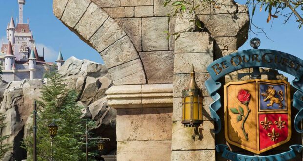 An ornate stone archway with a sign that reads "Be Our Guest Restaurant" invites you into Magic Kingdom Dining. In the background, a fantasy-style castle towers above rocky terrain and trees under a clear blue sky.