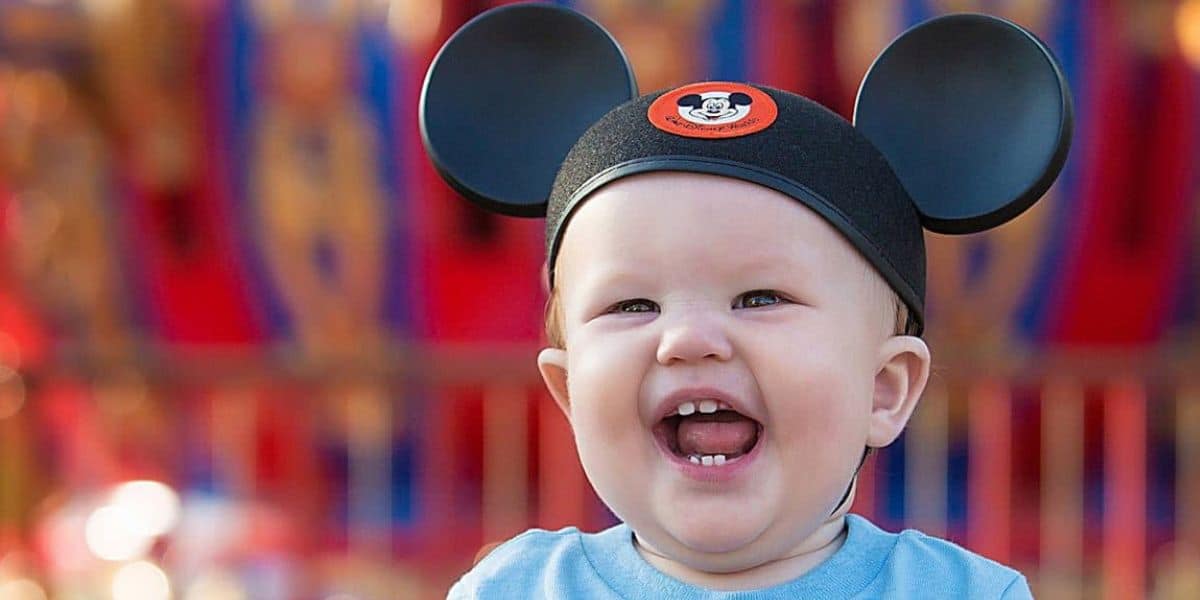 A baby wearing Mickey Mouse ears is smiling joyfully, capturing the magic of visiting Disney's EPCOT with a toddler. The colorful and blurred background evokes an amusement park setting, perfect for exploring Disney tips for moms on making memories with their little ones.