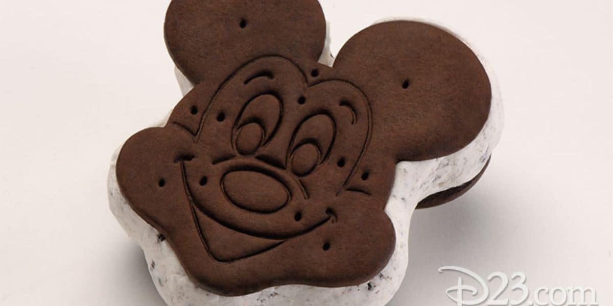 Enjoy a delightful Disney Treat with this Mickey Mouse-shaped ice cream sandwich. Featuring chocolate cookies and creamy vanilla ice cream, the face is intricately detailed on the top cookie. A "D23.com" logo graces the bottom right corner, celebrating iconic Disney Treats and Snacks.