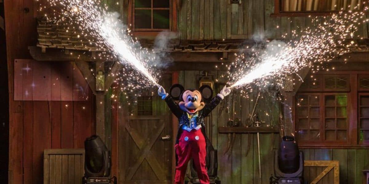 Mickey Mouse, dressed as a magician, stands with arms raised, shooting sparkling fireworks in front of a rustic wooden building. The scene captures the energetic and magical atmosphere reminiscent of a classic Disney Nighttime Show.
