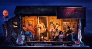 A vintage scene depicts a small gas station/store at night with an illuminated gas sign. People are sitting on the porch: one playing guitar, one on a stool, and another in a chef's uniform. An old Coca-Cola sign is visible, along with various other vintage advertisements. It's like stepping into Americana at Disney World.