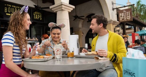 Three people sit at an outdoor restaurant table, smiling and enjoying a meal in a festive atmosphere. One wears mouse-ear headbands, hinting at Disney fun. In the background, a sign for "The Polite Pig" adds to the lively setting, making it an ideal spot for any Disney guest to unwind.