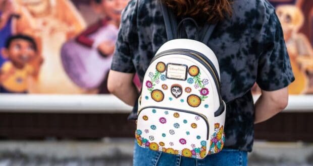 A person wearing a black tie-dye shirt stands with a trendy white backpack featuring colorful floral designs and a skull motif. The background is blurred, showcasing vibrant artwork with people and musical themes, reminiscent of the whimsical style found in New Disney Collections.