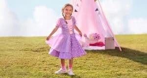 A young girl in a purple princess dress smiles and holds her skirt while standing on grass, celebrating Disney Princesses at home with a pink play tent and toys in the background on a sunny day.