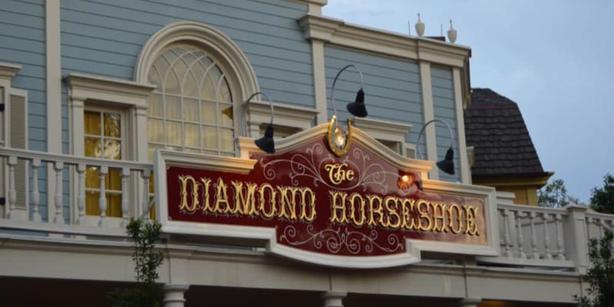 A two-story building with a light blue facade and white trim features an illuminated sign that reads "The Diamond Horseshoe" in decorative gold lettering on a red background. The sign is adorned with a horseshoe and several outdoor lights, marking one of the best places to eat at Magic Kingdom.