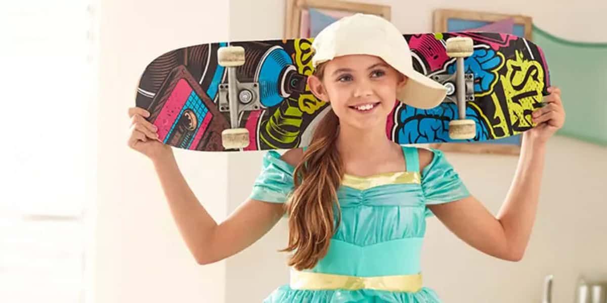 A smiling girl in a turquoise dress and white cap holds a colorful skateboard behind her neck, standing indoors—ready to celebrate Disney Princesses at home with style and fun.