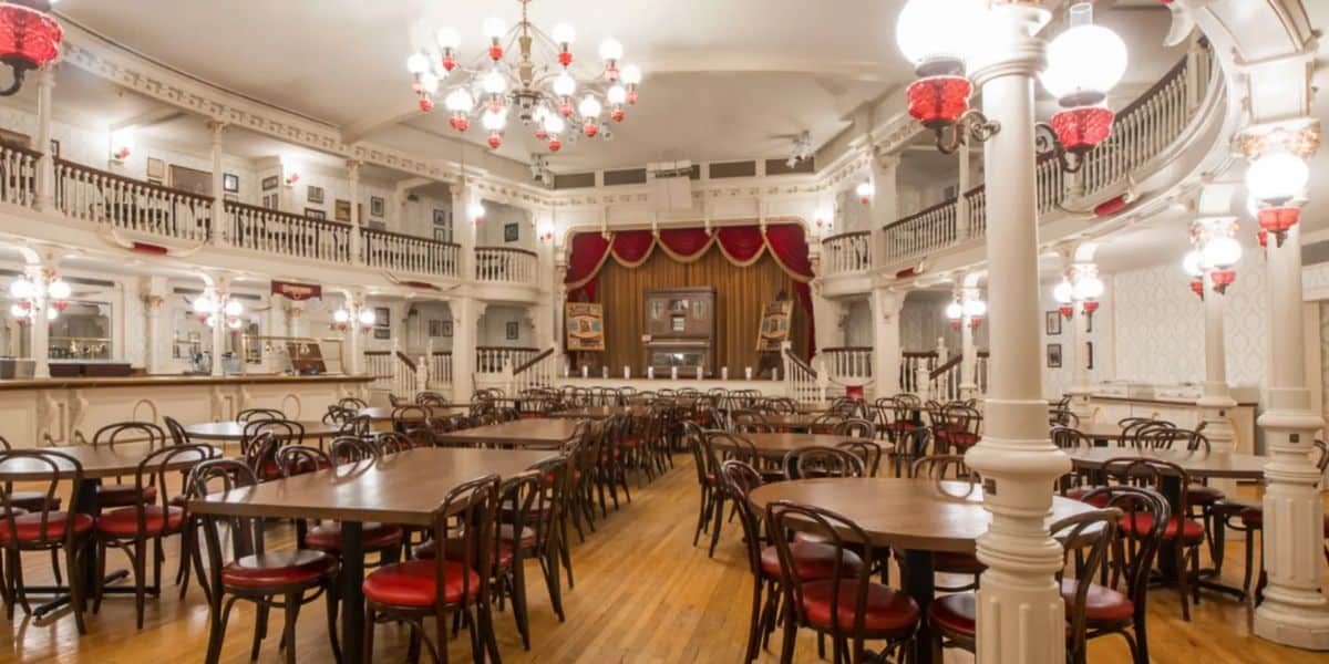 A vintage-style saloon, often hailed as one of the best dining spots at Disney World, boasts a spacious interior with wooden tables and red-cushioned chairs. Elegant white railings and columns adorn the room, while chandeliers and red-shaded lamps provide lighting. A stage with red curtains is at the far end.
