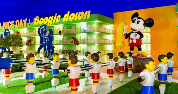 The vibrant exterior of the Disney Resort features large statues of a smiling mouse in a yellow outfit holding a phone, alongside a giant foosball table. Bright signs proudly display "Dance Da..." and "Boogie Down!" against the backdrop of a vivid blue sky.