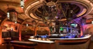 Futuristic bar with a circular design, illuminated by colored lights. The ceiling boasts a large, intricate apparatus with wires and lights above the counter area. Known for serving some of the best drinks at Disney World, it exudes an industrial, science fiction aesthetic.