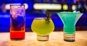 Three colorful cocktails on a bar counter: a tall glass with a red drink and ice, a round glass with a green drink garnished with rosemary, and Disney World's frosty blue signature cocktail in a wide-mouthed glass. A dim, blue-lit background enhances the scene.