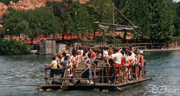 A group of people stand closely together on a wooden raft, floating on a calm body of water. Trees and a small structure reminiscent of the Magic Kingdom are visible in the background. The scene appears lively under the clear weather, with a whimsical air that hints at an adventure worthy of any bear's tale.