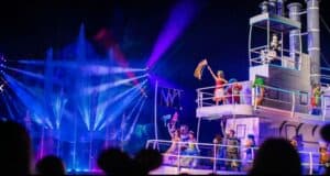 Performers in colorful costumes dance on a boat with vibrant lighting and water fountains in the background. The scene is lively, resembling a Disney Nighttime Performance, with neon blue and purple lights illuminating the stage, creating an energetic and festive atmosphere.