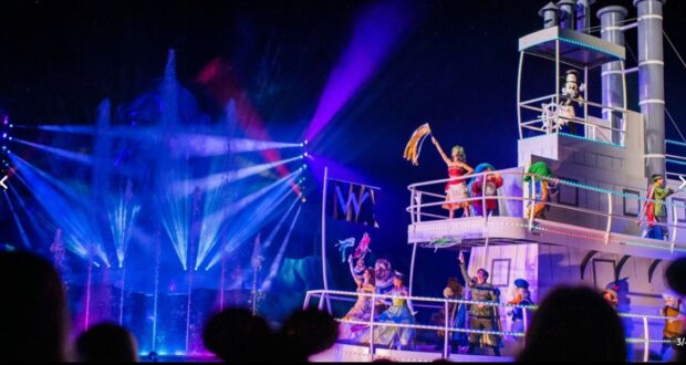 Performers in colorful costumes dance on a boat with vibrant lighting and water fountains in the background. The scene is lively, resembling a Disney Nighttime Performance, with neon blue and purple lights illuminating the stage, creating an energetic and festive atmosphere.