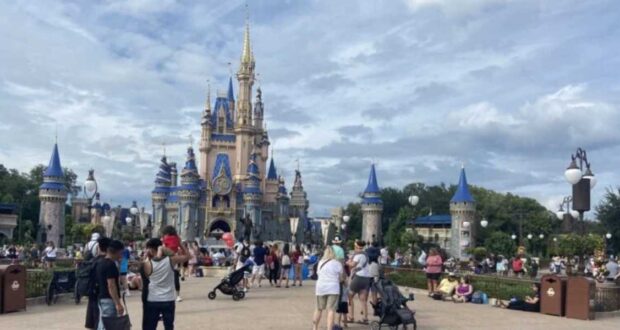 A bustling scene unfolds at a theme park with a large, ornate castle in the background. Visitors, mindful of the Disney World dress code, including families with strollers, gather joyfully in front, basking in the sunny day. The partly cloudy sky enhances the lively atmosphere.