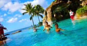 People enjoying swimming in a clear blue pool near the ocean at Disney Aulani. There are palm trees and a rocky structure with greenery in the background under a partly cloudy sky.