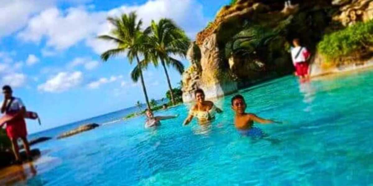 People enjoying swimming in a clear blue pool near the ocean at Disney Aulani. There are palm trees and a rocky structure with greenery in the background under a partly cloudy sky.