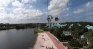 Experience a Disney World vacation with a scenic view of a cable car system gliding over a tree-lined resort area, featuring turquoise-roofed buildings. Marvel at the tranquil river on the left and enjoy the charming pathway below under a partly cloudy sky.