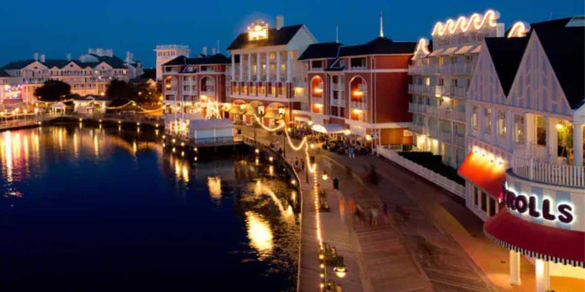 Nighttime scene of a lively waterfront boardwalk with colorful buildings, bright lights reflecting on the water, and people strolling along the path. The area has a festive atmosphere, featuring various shops and restaurants.