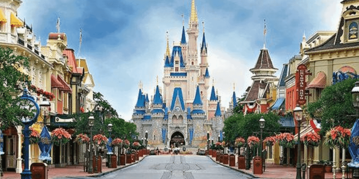 A view of a vibrant theme park street leading to a large, enchanting castle with blue and gold spires. Decorative buildings line the path, adorned with flowers. The sky is clear and bright, enhancing the whimsical atmosphere.