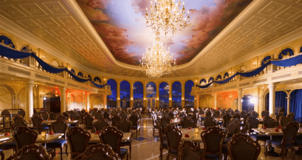 Elegant, grand dining hall with ornate chandeliers and a painted ceiling, offering the best dining at Magic Kingdom park. Rows of lavish tables and chairs fill the space. Blue drapery and arched windows enhance the opulent, palace-like atmosphere.