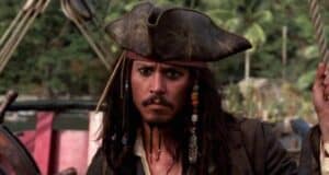 A man dressed as a pirate with long hair, a tricorn hat, beads in his hair, and a serious expression stands on a ship with greenery in the background, reminiscent of Johnny Depp's iconic portrayal.