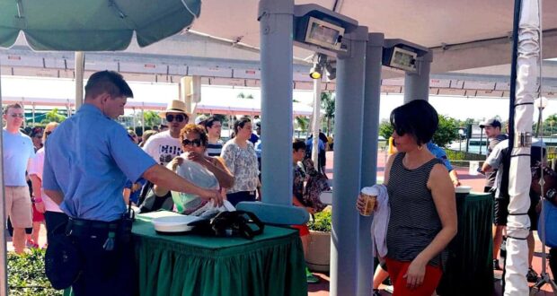 A security guard checks a bag at an outdoor checkpoint as people wait in line; a woman holding a drink stands by, and several people are in the background under a canopy on a sunny day.