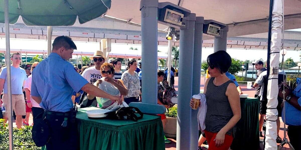 A security guard checks a bag at an outdoor checkpoint as people wait in line; a woman holding a drink stands by, and several people are in the background under a canopy on a sunny day.