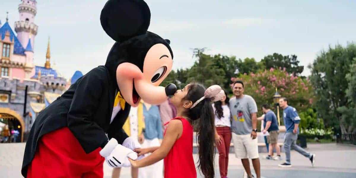 A young girl in a red dress is joyfully interacting with Mickey Mouse in front of a castle at Disneyland. Despite the cheerful scene, whispers of a recent blackout float through the crowd, yet people continue to walk and smile under the clear skies and green trees.