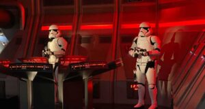 Two Stormtroopers in white armor stand guard inside a spaceship control room with red lighting, standing beside futuristic control panels glowing with red lights.