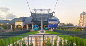 Two gondolas on a cable car system are stationed at a platform near a building with a sign reading "Riviera Resort." The sky is partly cloudy, and there's a modern building in the background. A fence and greenery are in the foreground.
