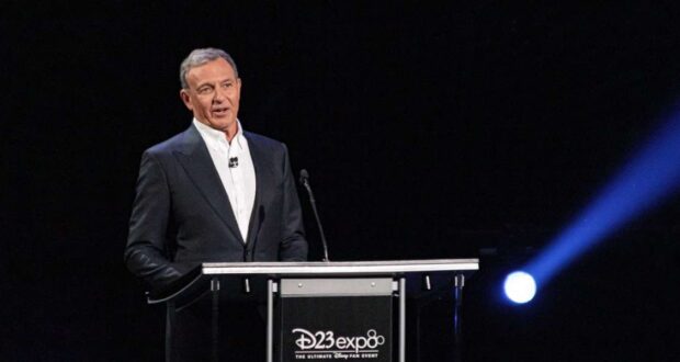 A man in a dark suit speaks at a podium with a microphone, with the "D23 Expo" logo on the front, against a dark background with a bright spotlight.