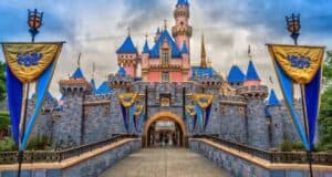 Disneyland’s Sleeping Beauty Castle with blue rooftops and pink walls, framed by blue and gold banners, sits at the end of a stone walkway under a cloudy sky.