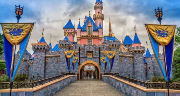 Disneyland’s Sleeping Beauty Castle with blue rooftops and pink walls, framed by blue and gold banners, sits at the end of a stone walkway under a cloudy sky.