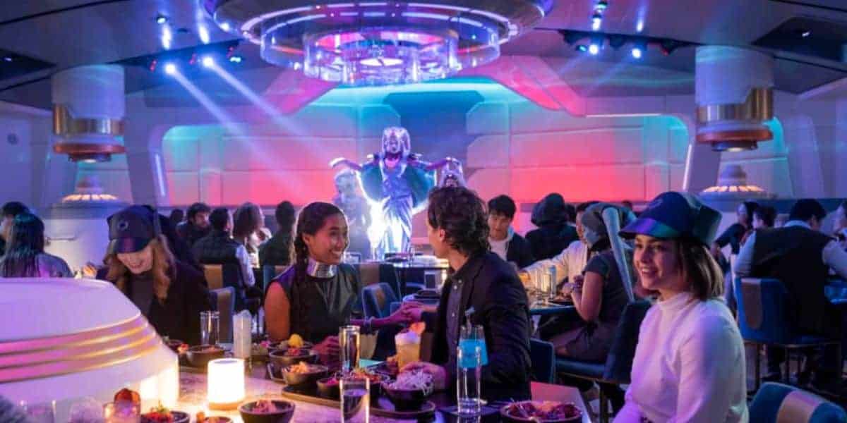 People dine in a futuristic, space-themed restaurant with colorful lighting. A performer in an elaborate alien costume entertains guests at the front of the room. Tables are filled with food and drinks.