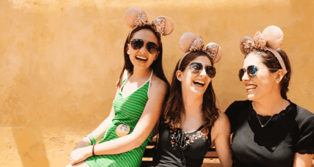 Three women wearing Disney-inspired ear headbands and sunglasses sit on a bench, laughing together against a yellow wall. The woman on the left is in a green dress, the middle in a black top, and the right in a dark dress—bringing cheer even during any Disneyland blackout.