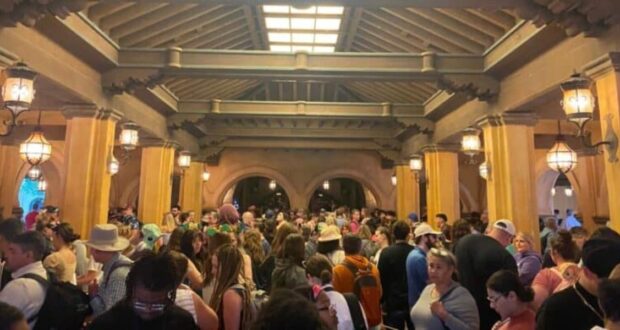 A large crowd gathers under a covered walkway adorned with wooden beams and lanterns. The bustling atmosphere is filled with excitement as people wait, some clutching their phones accessing Disney Genie and wearing hats. Numerous arches create a magical backdrop in the distance.