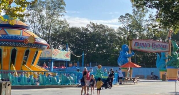 People stroll through a vibrant section of Animal Kingdom, featuring a spaceship-themed ride on the left and dinosaur decorations holding a "Dino-Rama!" sign on the right. In the background, trees frame the scene against a clear sky.