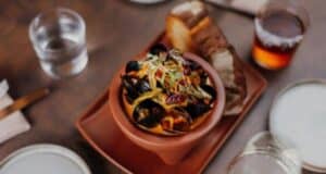 A bowl of mussels with vegetables and herbs sits on a wooden table, reminiscent of the vibrant flavors found at Downtown Disney restaurants. Accompanied by slices of bread and surrounded by glasses of water and an amber-colored beverage, it promises a delightful dining experience.