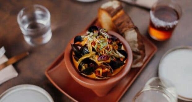 A bowl of mussels with vegetables and herbs sits on a wooden table, reminiscent of the vibrant flavors found at Downtown Disney restaurants. Accompanied by slices of bread and surrounded by glasses of water and an amber-colored beverage, it promises a delightful dining experience.