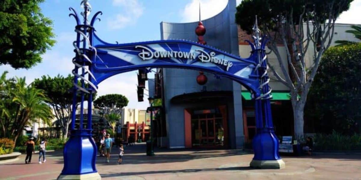 The entrance archway to Downtown Disney at Disneyland Resort showcases a blue and silver geometric design, surrounded by lush trees and bustling visitors in the background, all under a clear sky.