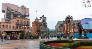 In this Disney Park scene, a statue of a man holding a mouse's hand graces the foreground. Surrounding buildings, including "The Hollywood Tower Hotel," showcase art deco architecture as people stroll along the wet pathways under a cloudy sky.