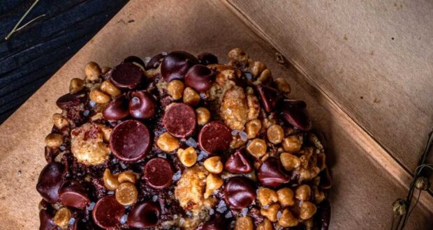 A close-up of a richly textured Gideons cookie topped with an assortment of chocolate chips and caramel pieces. The cookie is displayed on a wooden surface, with a dark background that subtly shows some dried twigs.